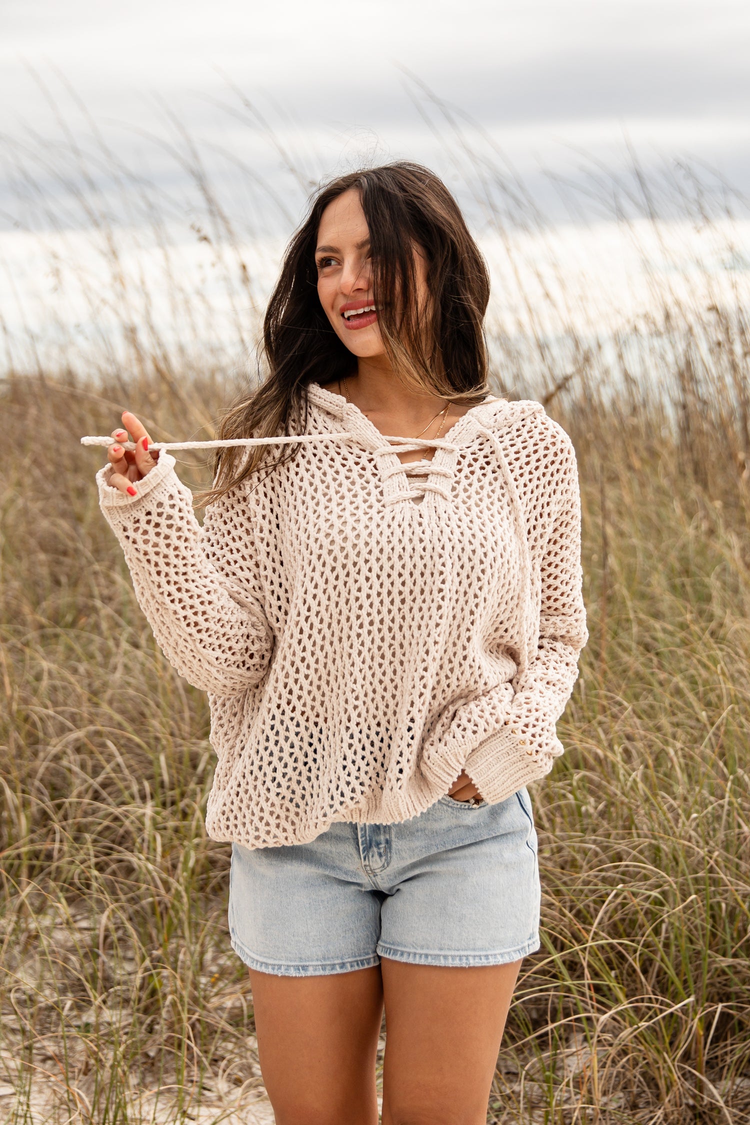 Woman wearing a beige crochet sweater and denim shorts standing in a field.