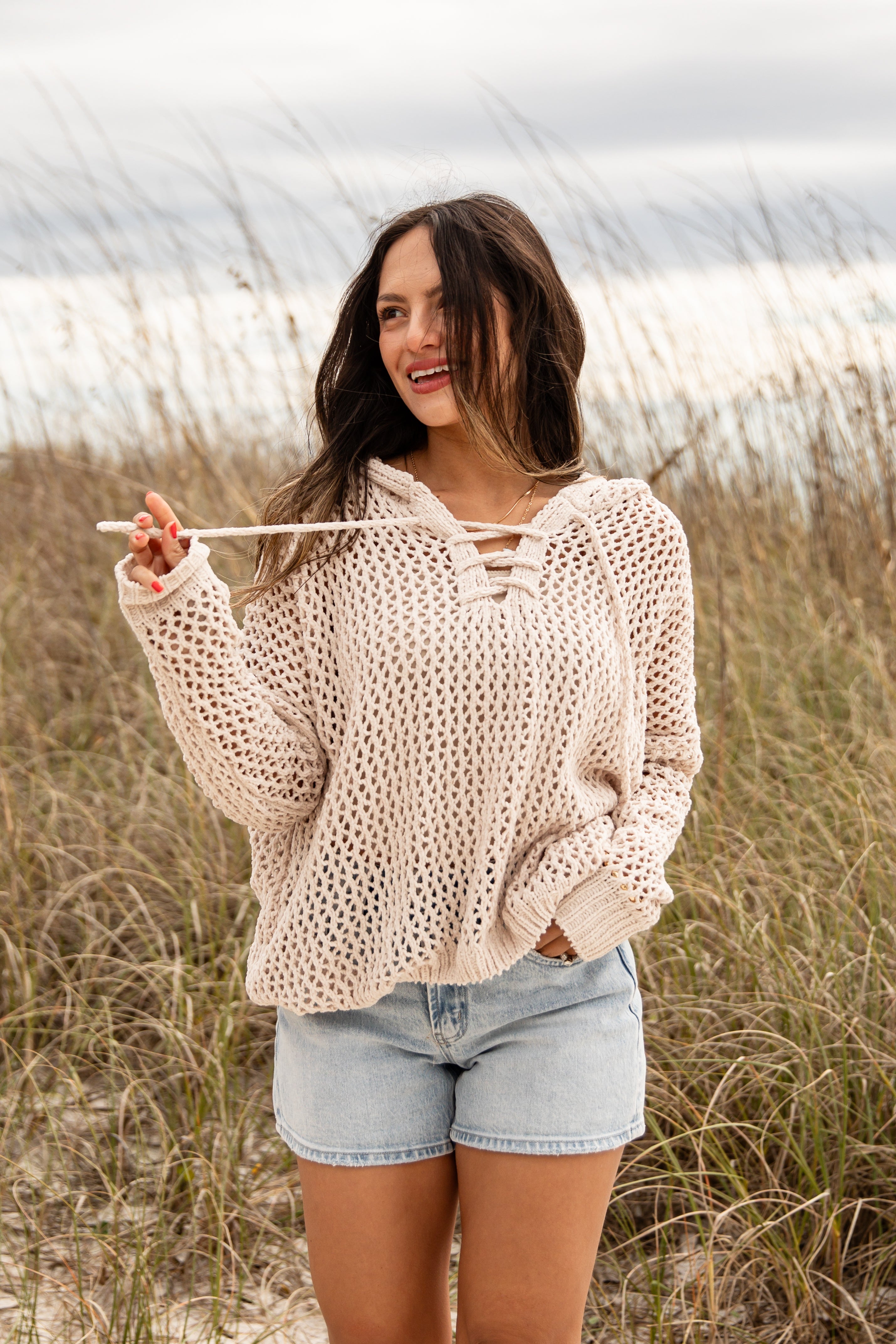 Woman wearing a beige crochet sweater and denim shorts standing in a field.
