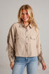 A smiling blonde woman wearing a cropped beige fringed button-up shirt with blue jeans, standing against a light gray brick wall.
