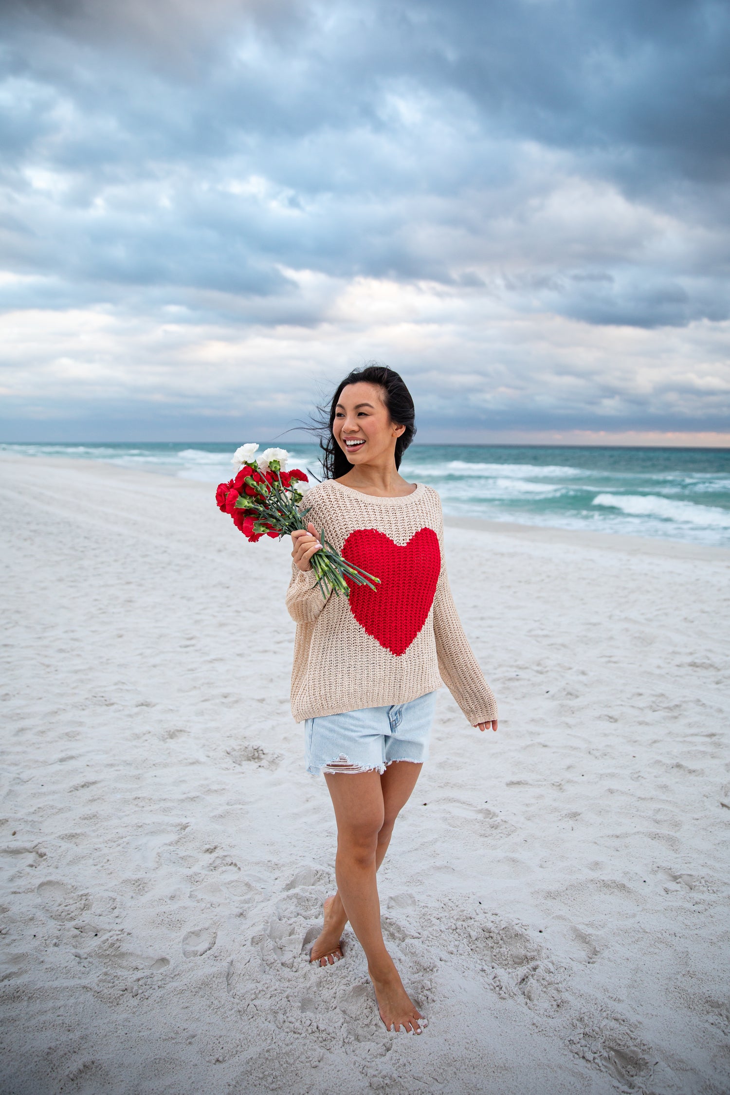 Woman on a beach holding flowers and wearing a sweater with a heart design.