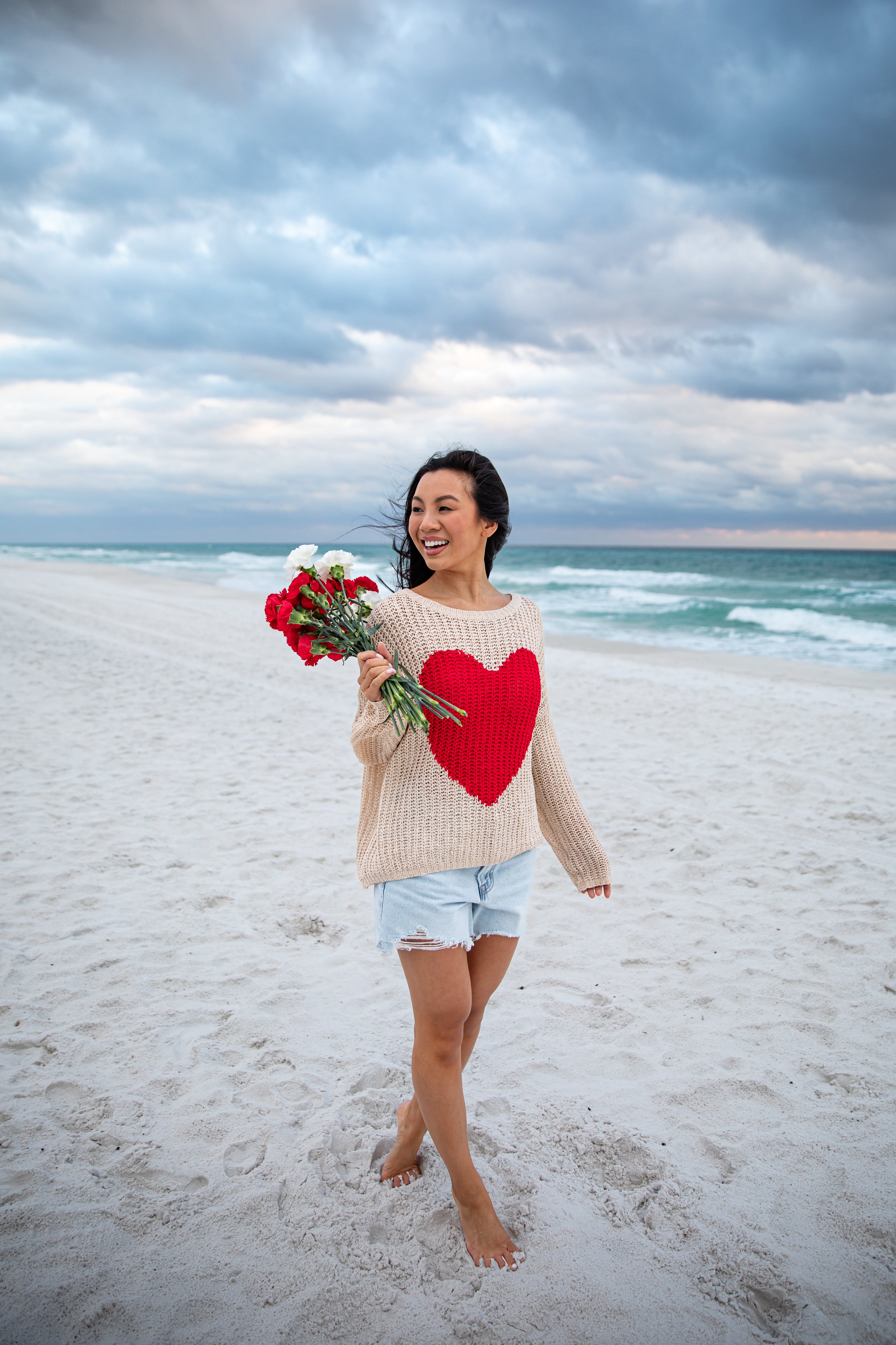 Woman on a beach holding flowers and wearing a sweater with a heart design.