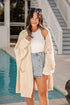 Woman wearing a beige cardigan, white top, and denim skirt by a pool.