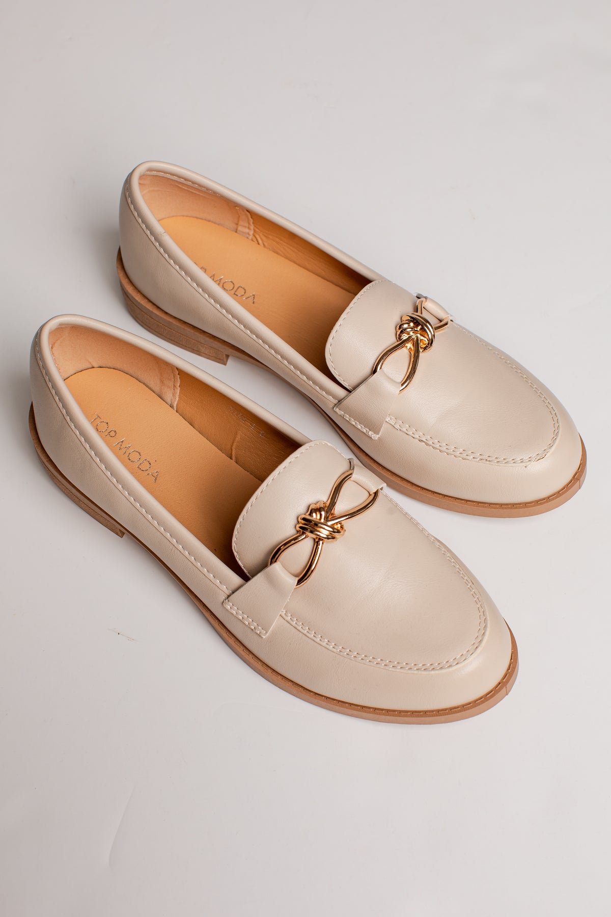 A pair of light beige leather penny loafers with a gold-tone metal bow detail across the vamp, displayed on a light gray background.