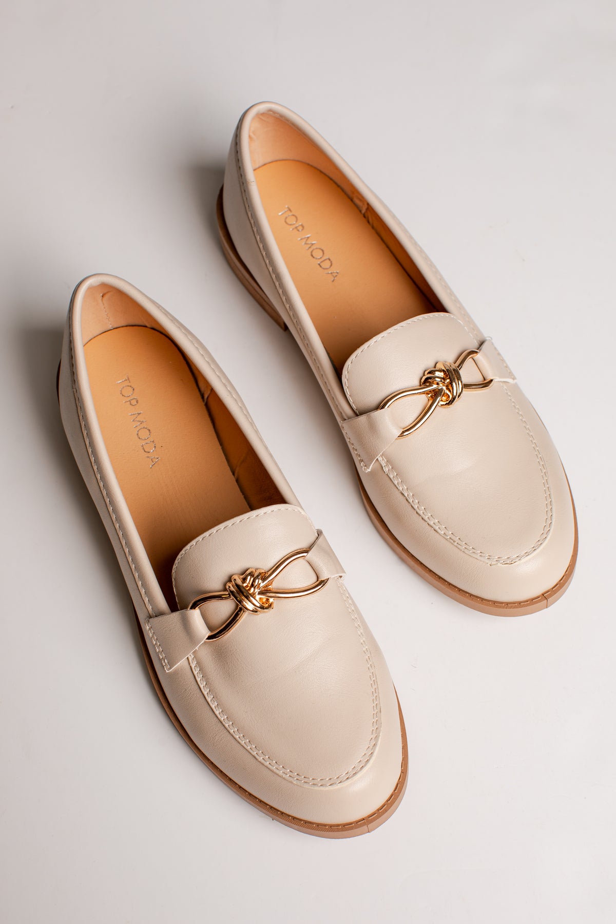 A pair of cream-colored leather penny loafers with a gold bow detail on the vamp, displayed on a light gray background.