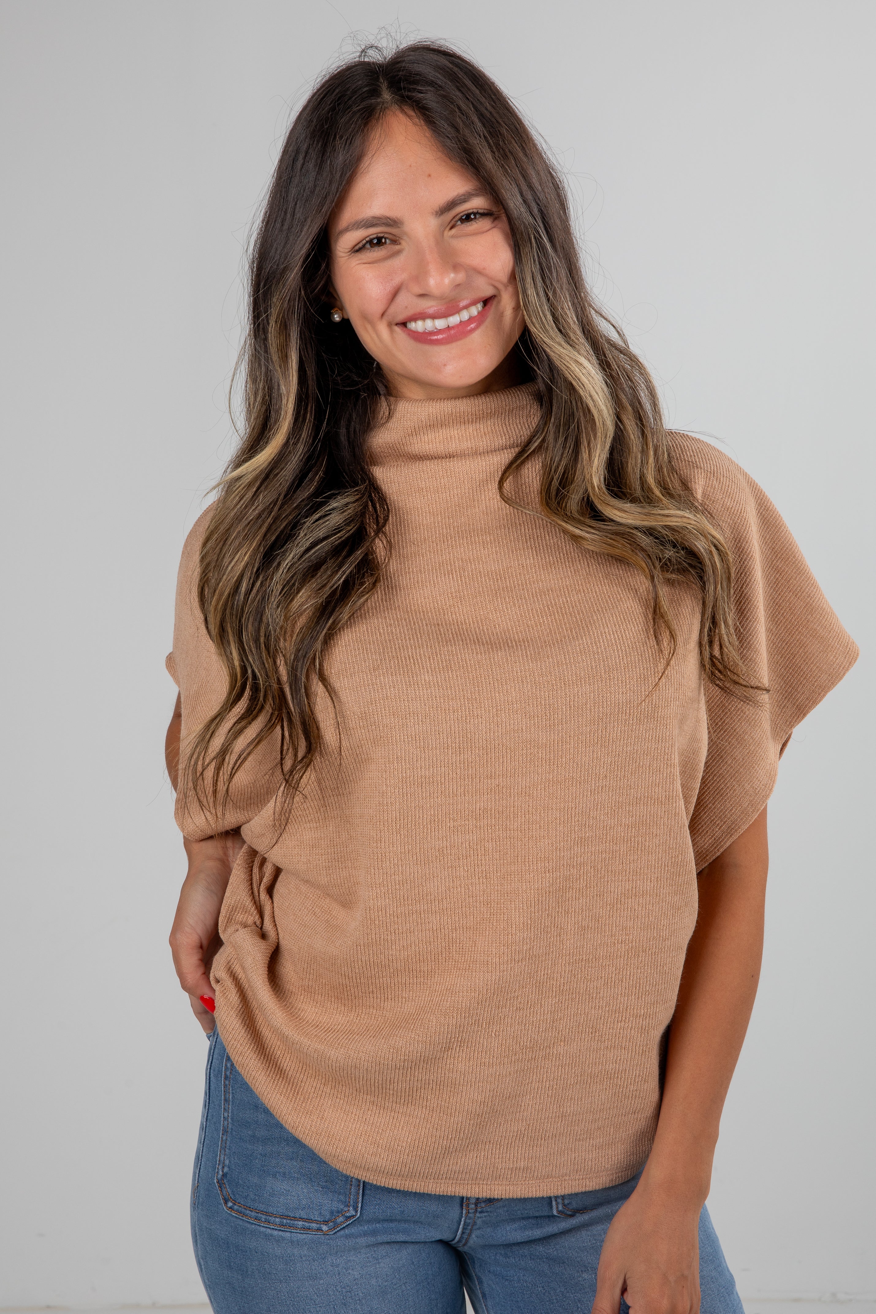 Woman wearing a brown turtleneck sweater and blue jeans against a plain background