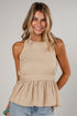 A smiling woman wearing a sleeveless beige peplum top and light blue jeans, photographed against a plain light gray studio background.