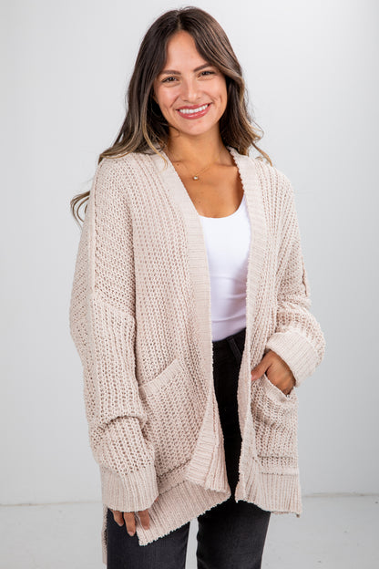 Woman wearing a beige knitted cardigan over a white top and black pants on a white background