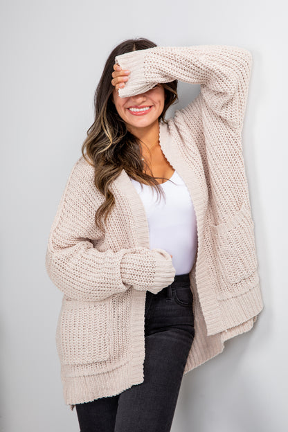 Woman wearing a beige chunky knit cardigan over a white top and black pants on a light gray background