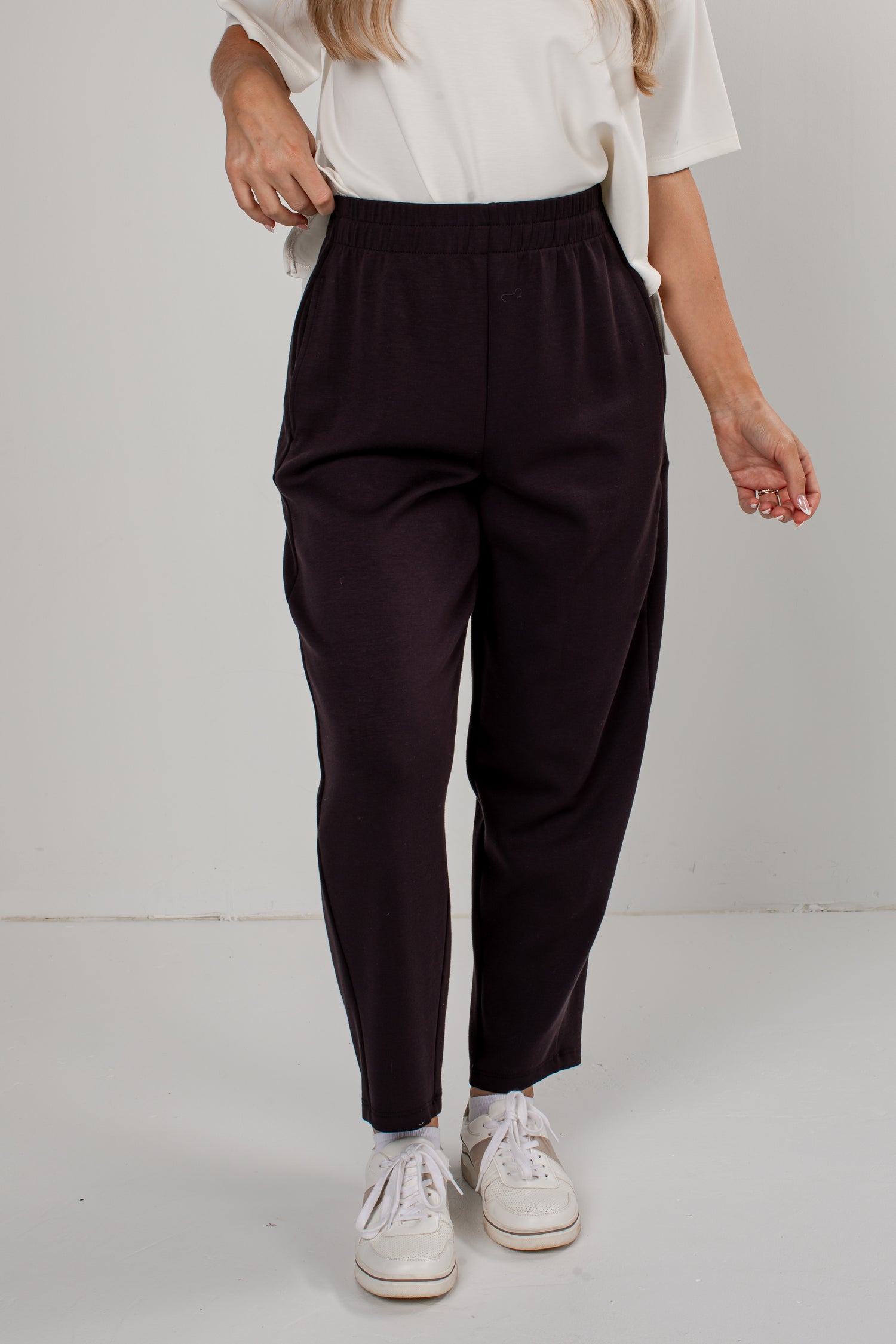 A person wearing dark black tapered pants with an elastic waistband, styled with a white top and white sneakers against a plain light gray studio background.