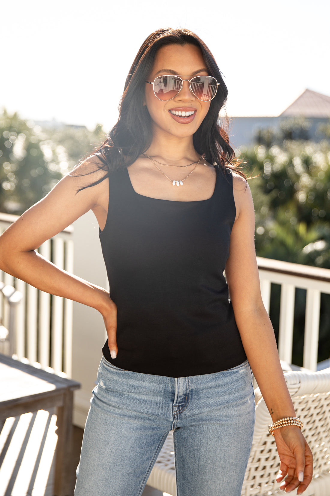 Woman wearing a black tank top and blue jeans standing on a balcony with a blurred background