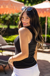 Woman in a black tank top and white pants standing outdoors with greenery and orange umbrellas in the background.