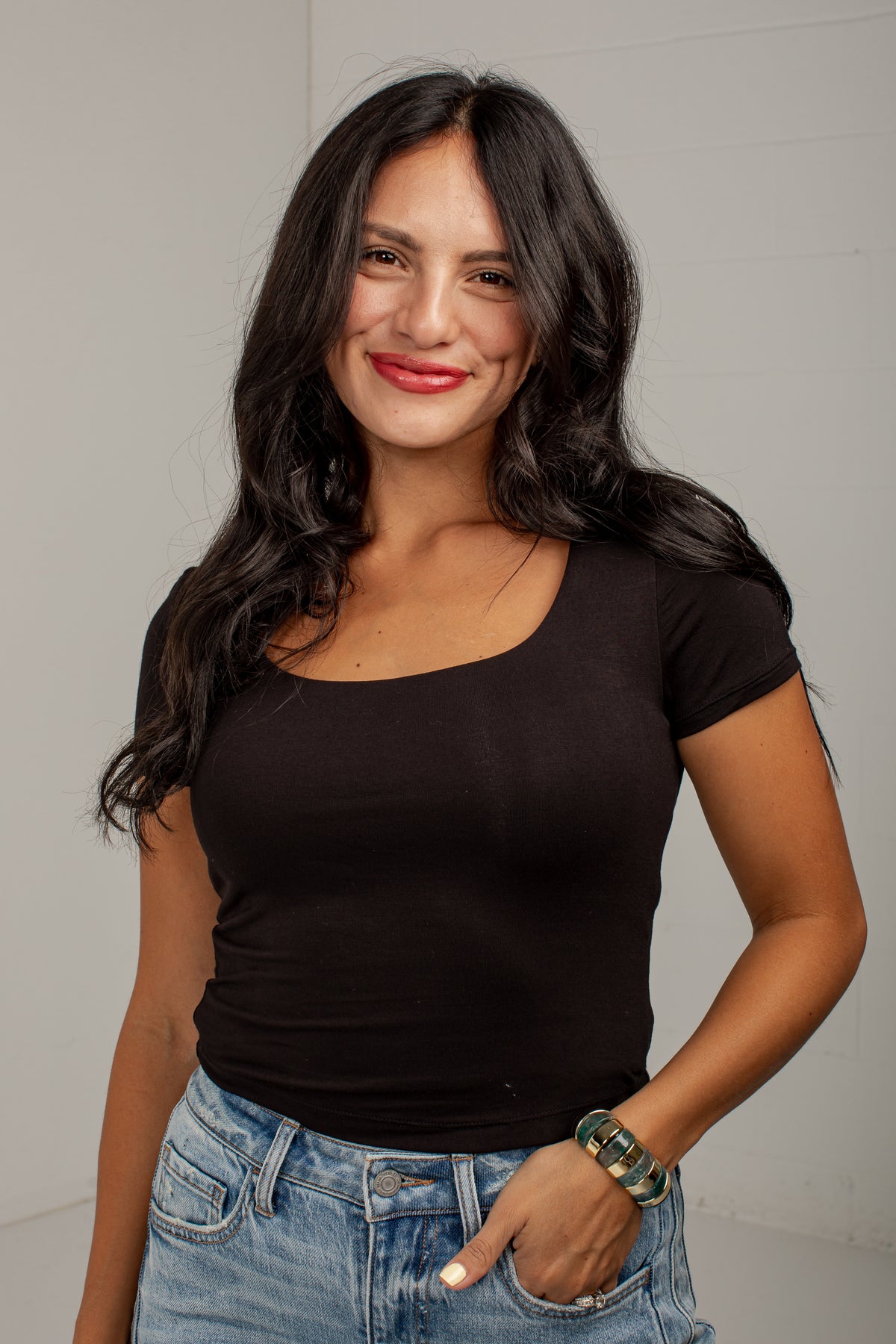 A woman with long dark hair wearing a fitted black scoop-neck short-sleeve top and light-wash blue jeans, standing against a light gray wall.