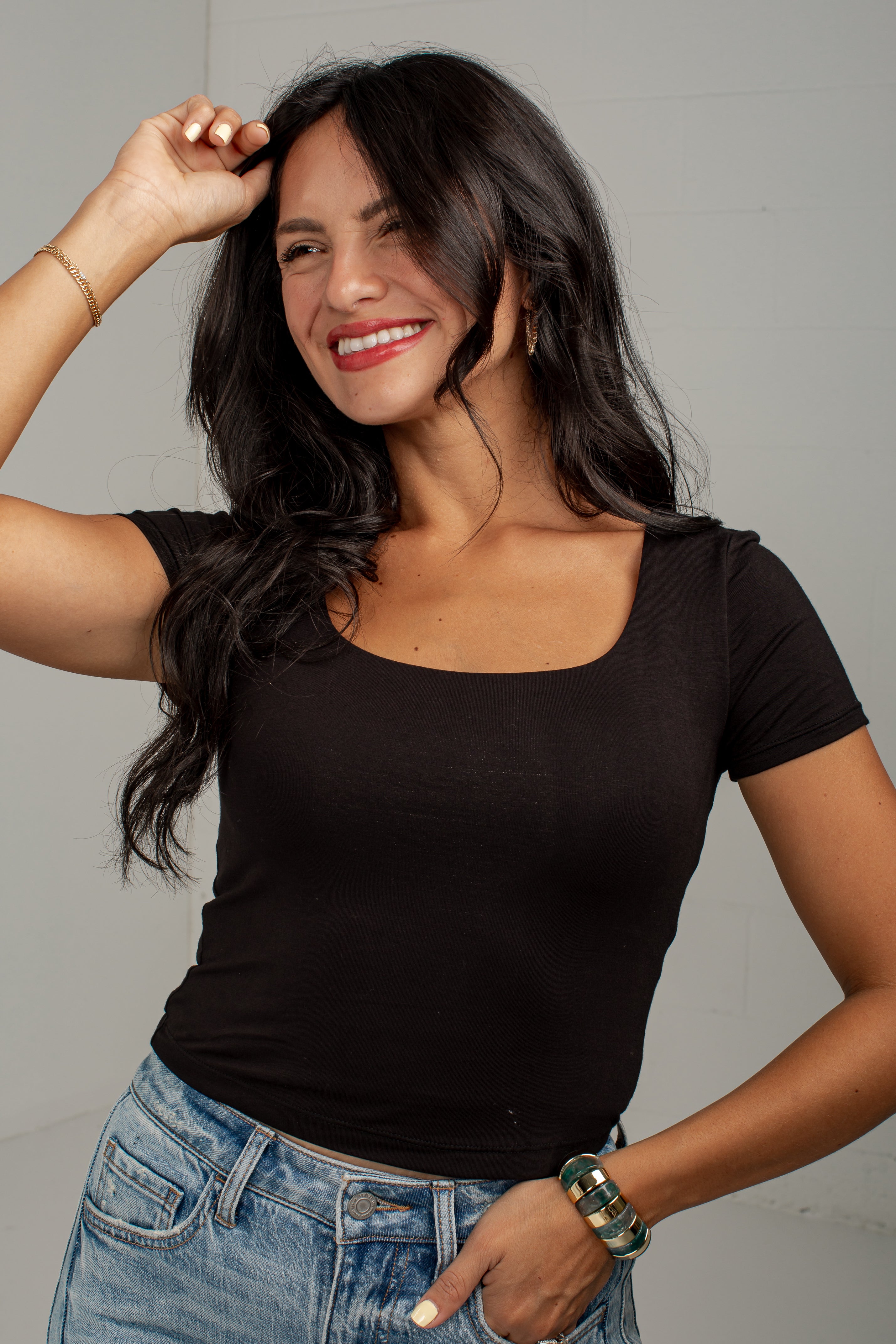 A woman with long dark hair wearing a fitted black scoop-neck short-sleeve t-shirt and light blue jeans, posing against a light gray wall.