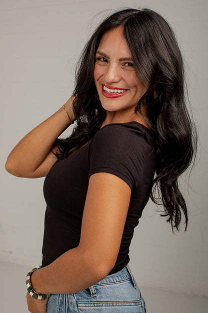 A woman with long, wavy black hair smiling and looking over her shoulder, wearing a fitted black short-sleeve top and light blue jeans against a plain light gray studio background.