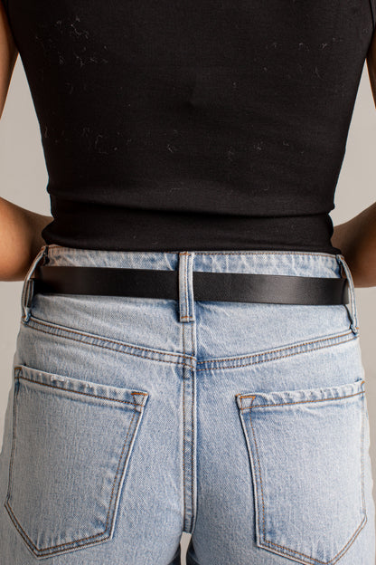 Close-up back view of a person wearing a black ribbed tank top, light-wash blue jeans, and a slim black belt against a plain light gray background.