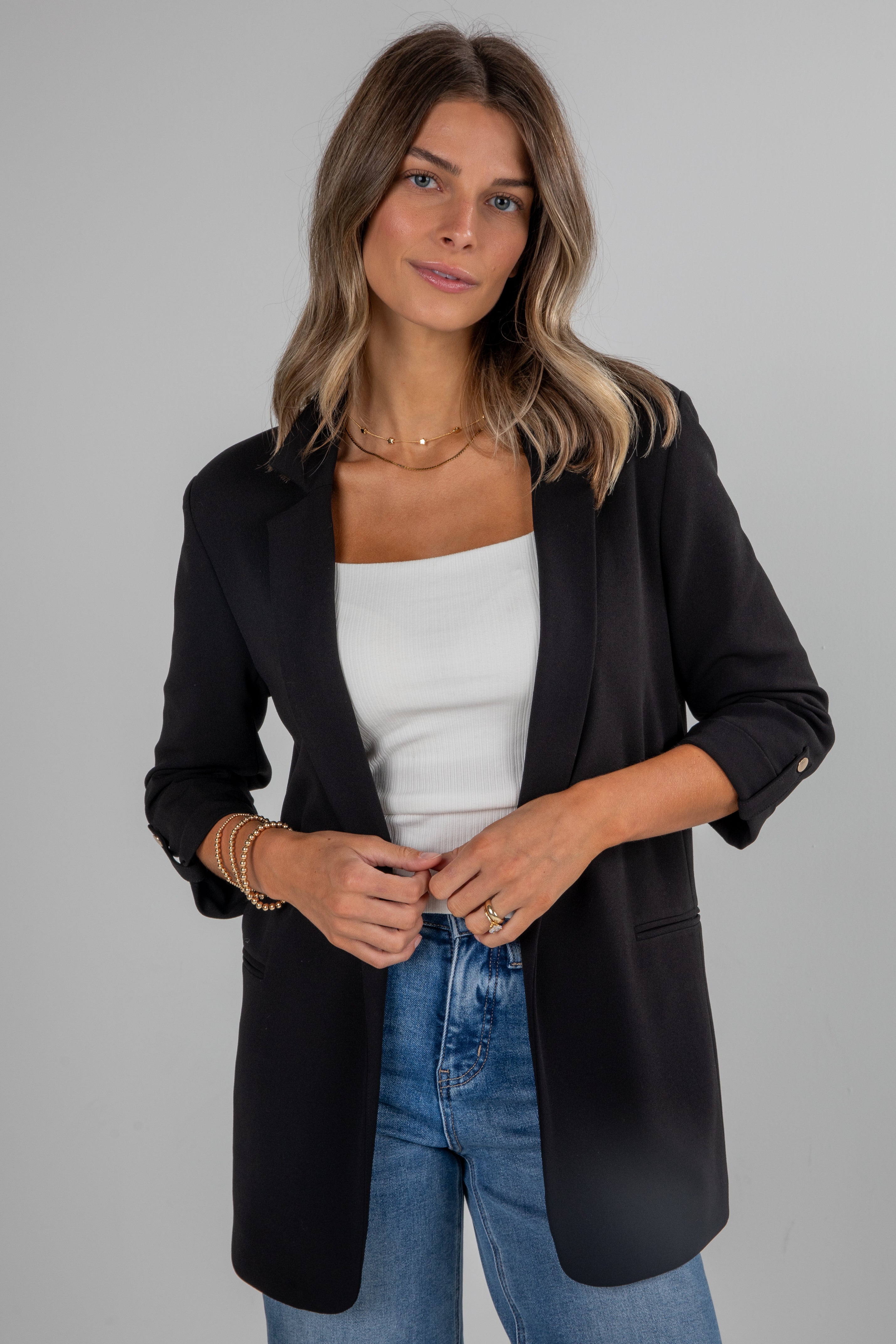 Woman wearing a black blazer over a white top and blue jeans on a gray background