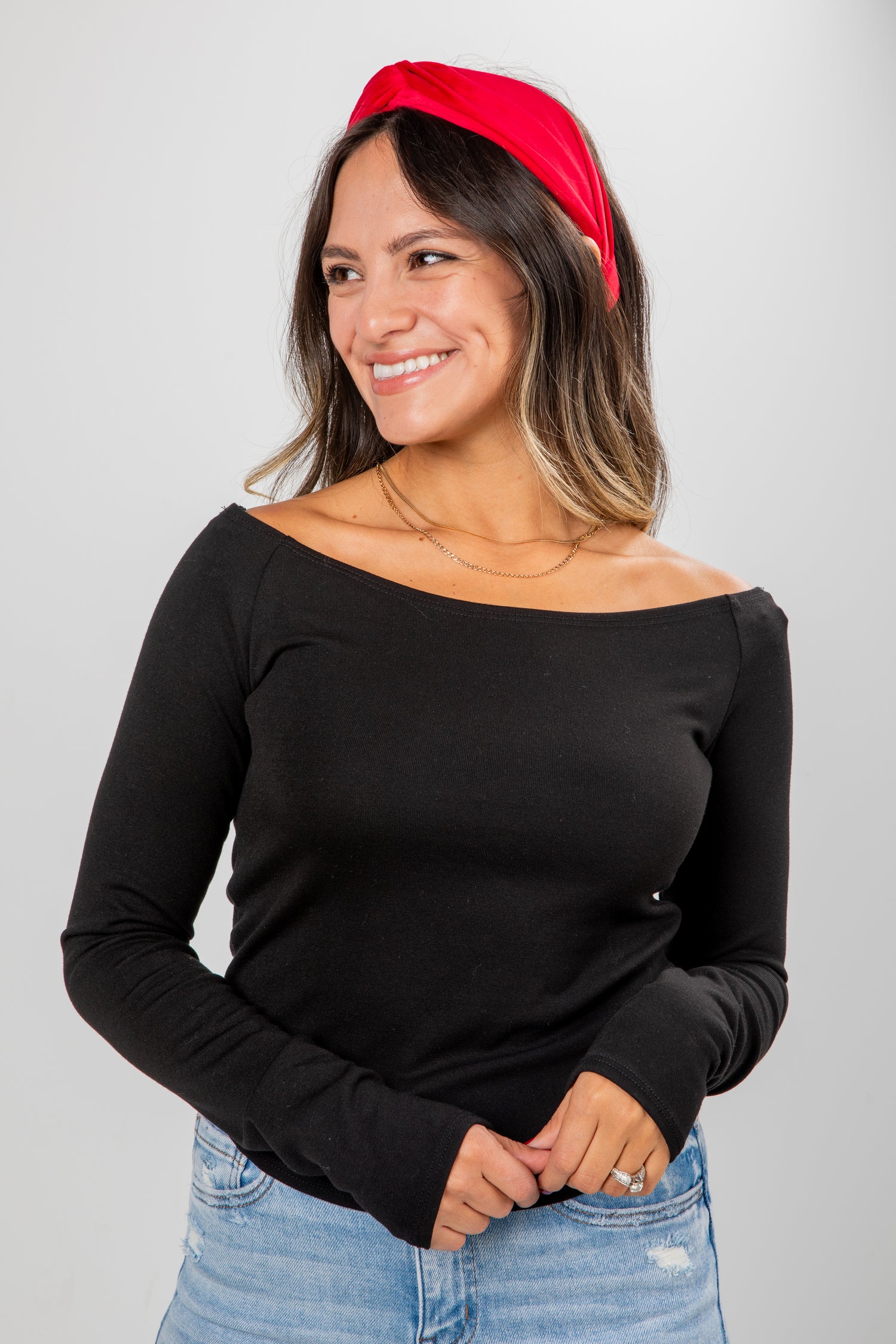 Woman wearing a black off-shoulder top and blue jeans with a red headband against a gray background
