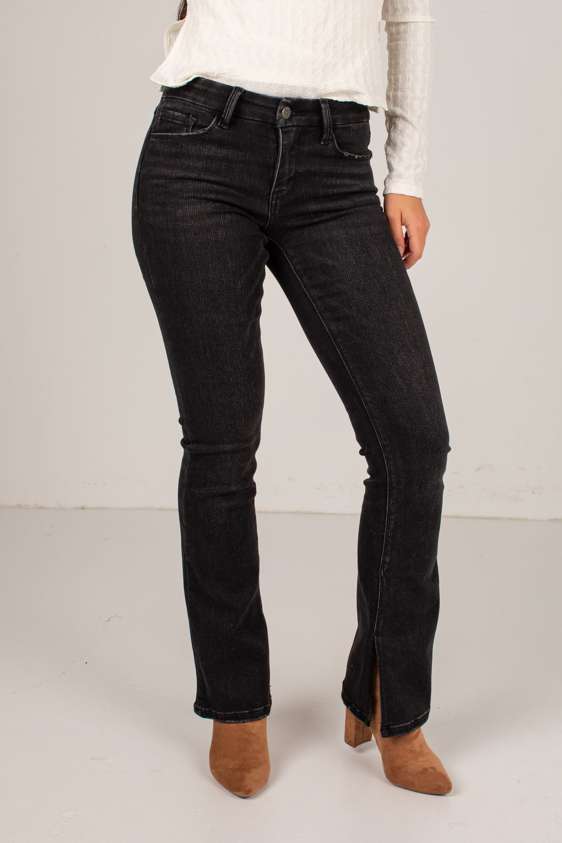 A model wearing black denim jeans with a subtle flare at the ankles, styled with a white ribbed long-sleeve top and brown suede ankle boots against a plain light gray studio background.
