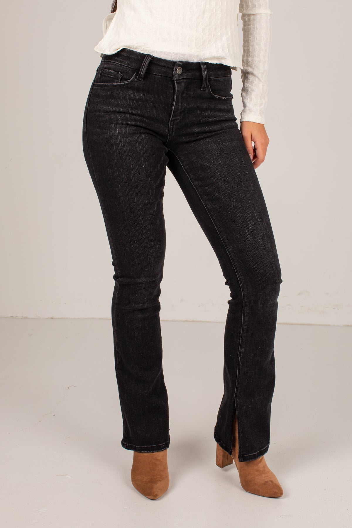 A model wearing black denim jeans with a subtle flare at the ankles, styled with a white ribbed long-sleeve top and brown suede ankle boots against a plain light gray studio background.