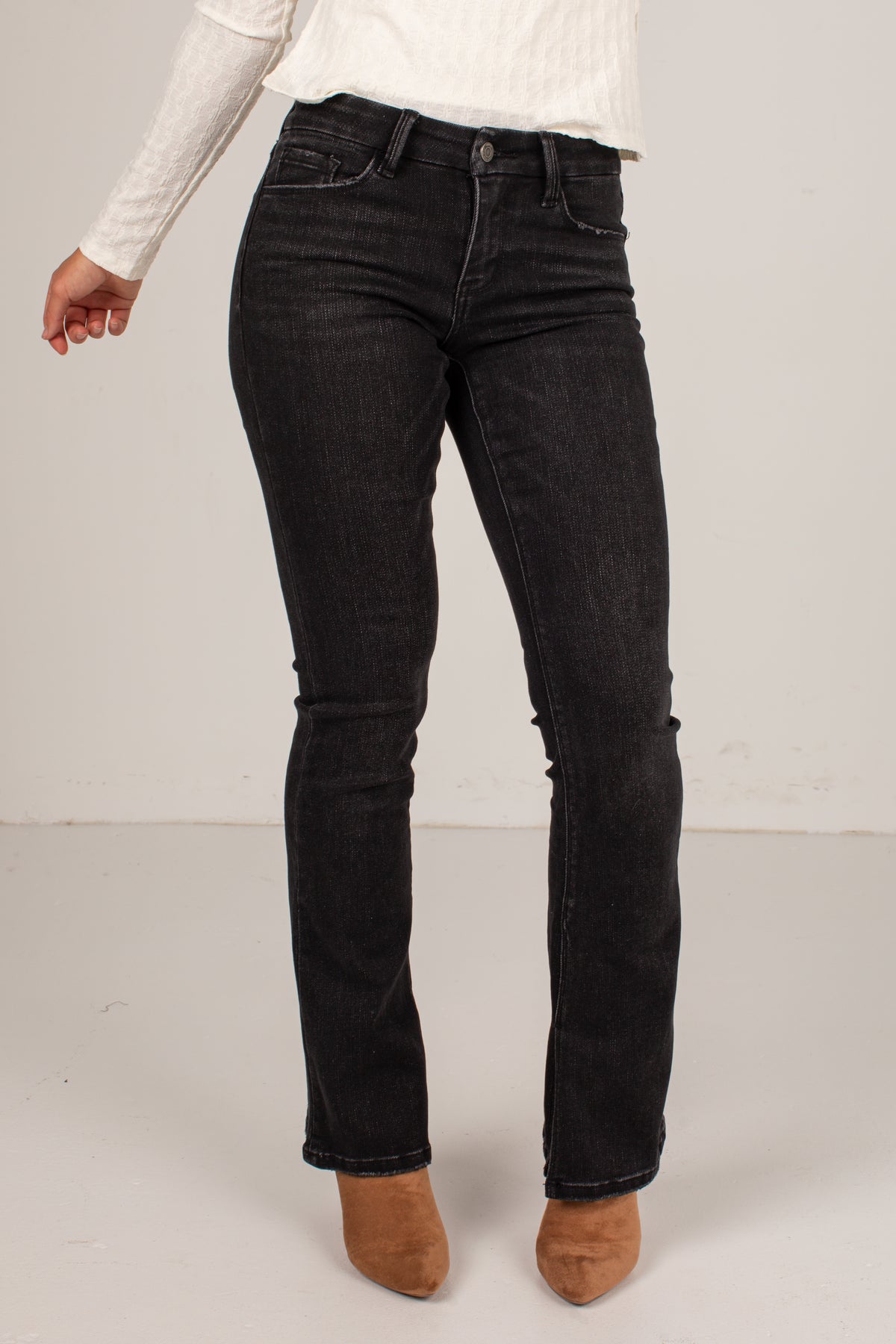 A person wearing black denim jeans, a white ribbed long-sleeve top, and brown suede ankle boots, shown from the waist down against a plain light gray studio background.