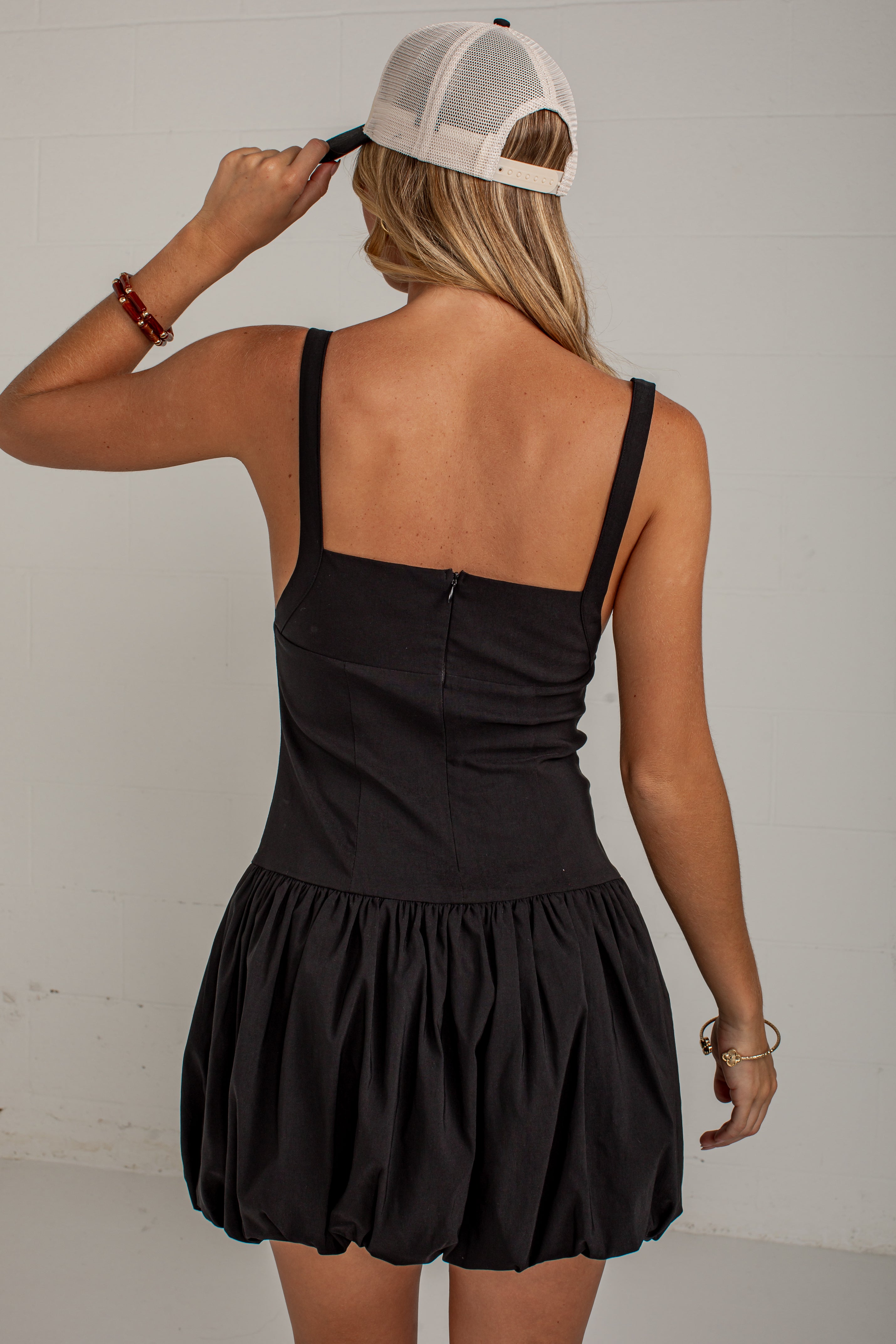 A woman shown from the back wearing a black sleeveless mini dress with thin straps and a gathered ruffled skirt, paired with a white trucker hat. She is standing against a light gray wall.