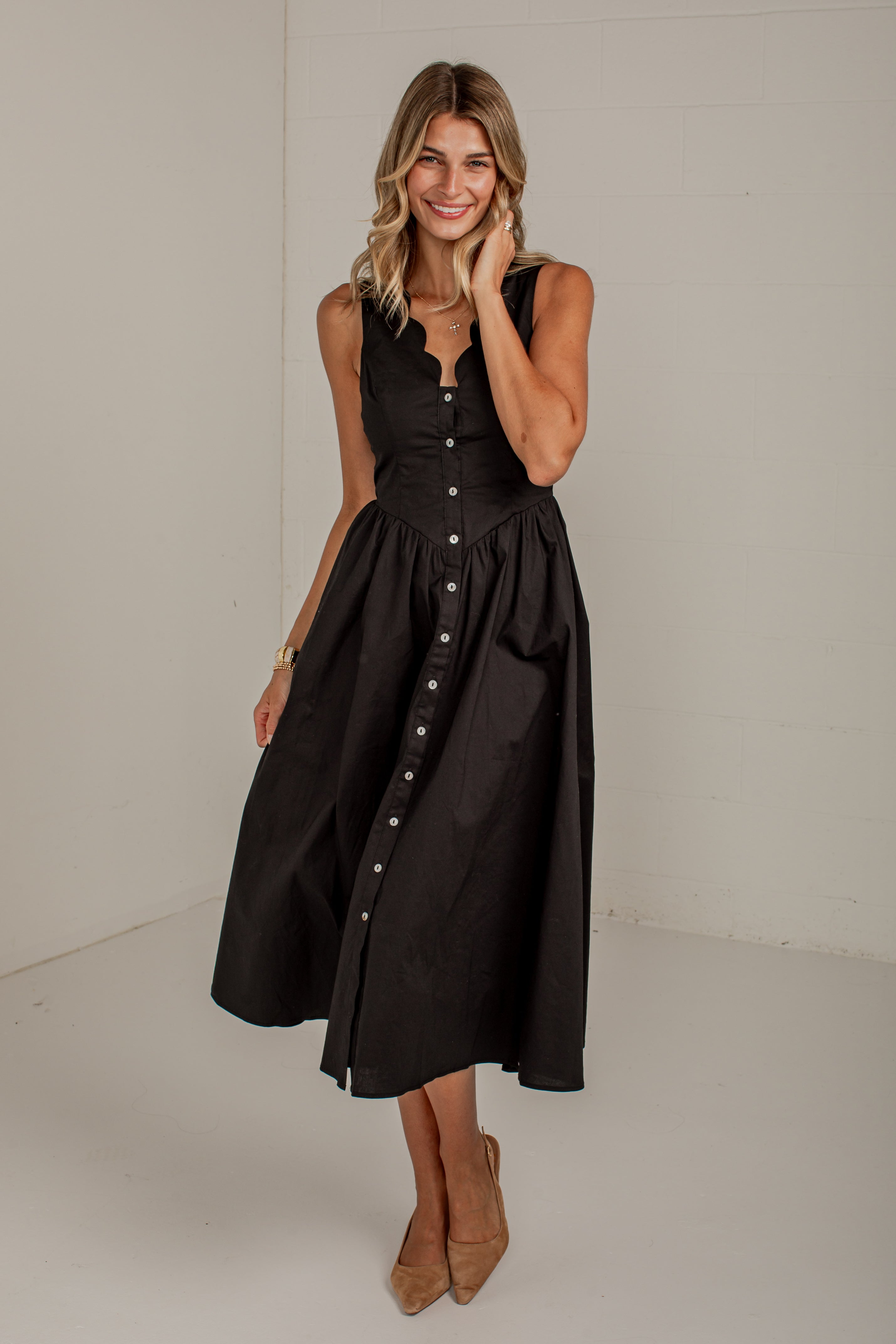 A woman modeling a sleeveless black midi dress with a buttoned front and gathered skirt, standing against a plain light gray studio background.