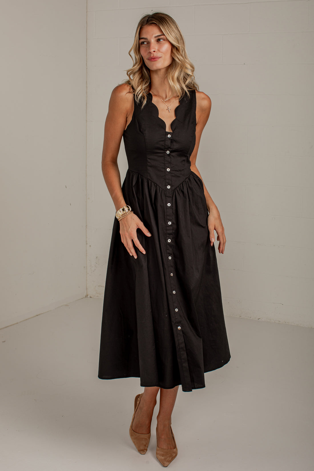 A woman modeling a sleeveless black midi dress with a scalloped V-neckline, buttoned bodice, and a flared skirt, standing against a plain off-white studio wall.