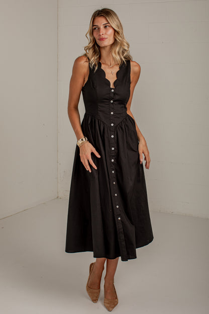 A woman modeling a sleeveless black midi dress with a scalloped V-neckline, buttoned bodice, and a flared skirt, standing against a plain off-white studio wall.
