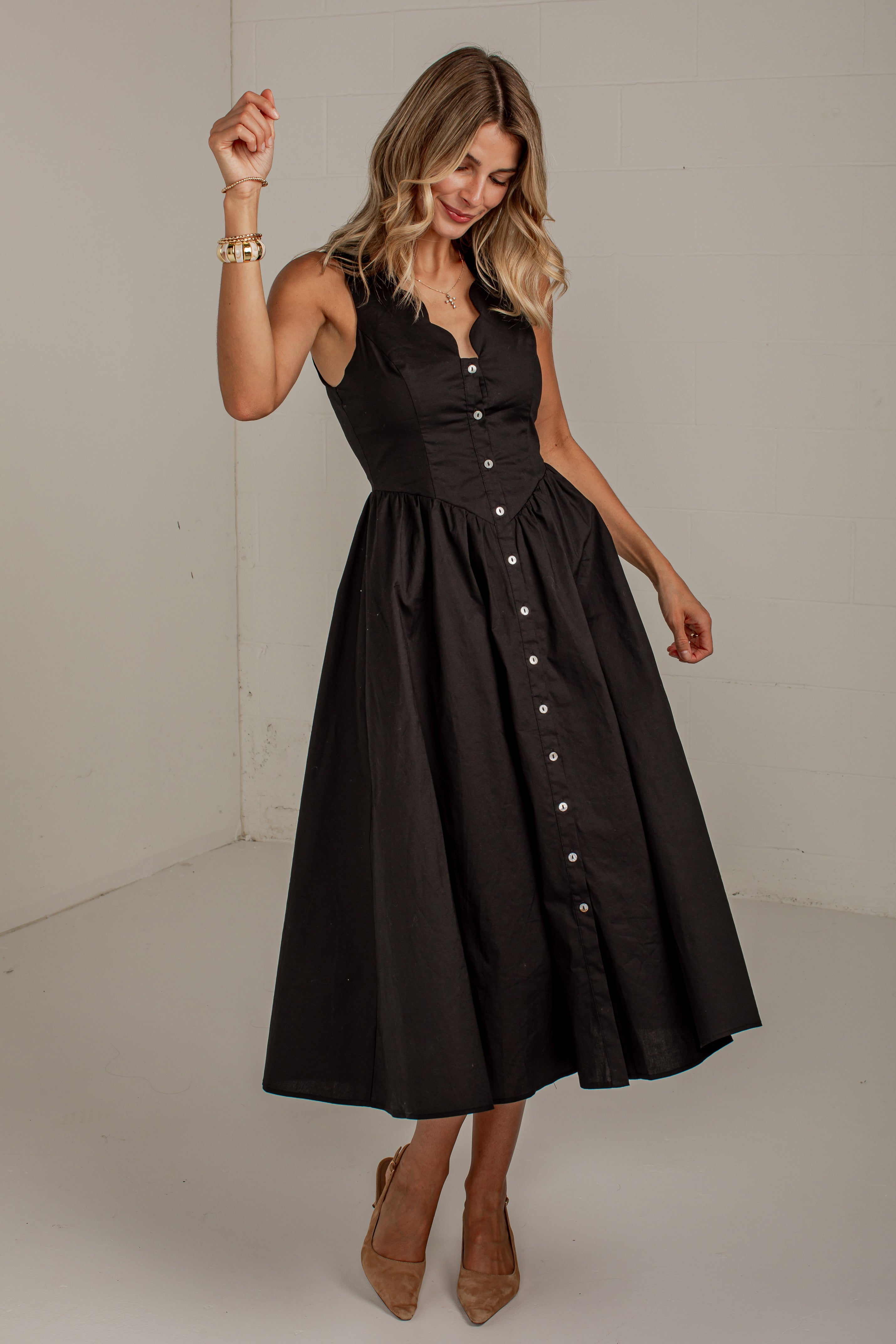 A woman modeling a sleeveless black midi dress with a structured corset-style bodice, flared skirt, and front button placket, photographed in a plain indoor studio setting.