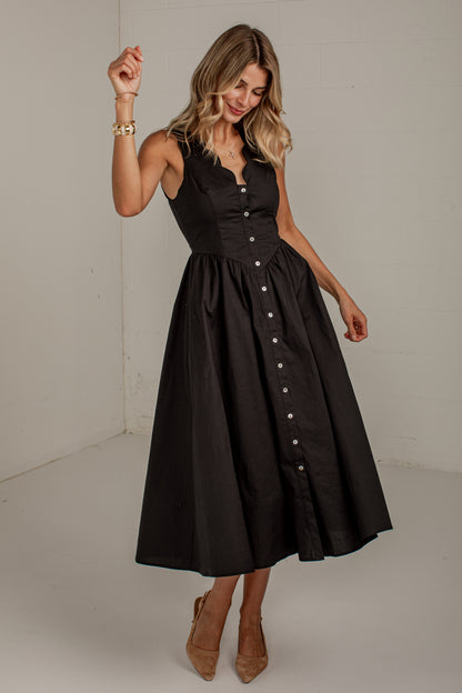 A woman modeling a sleeveless black midi dress with a structured corset-style bodice, flared skirt, and front button placket, photographed in a plain indoor studio setting.