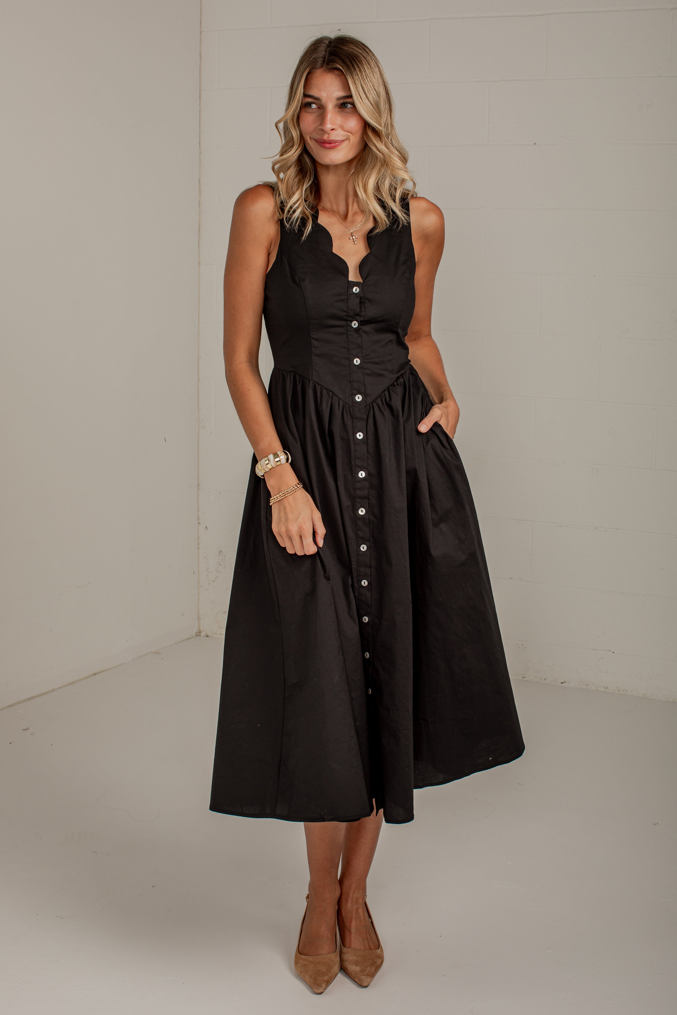 A woman modeling a sleeveless black midi dress with a scalloped V-neckline, button-front bodice, and a full skirt with side pockets, standing against a plain off-white studio wall.