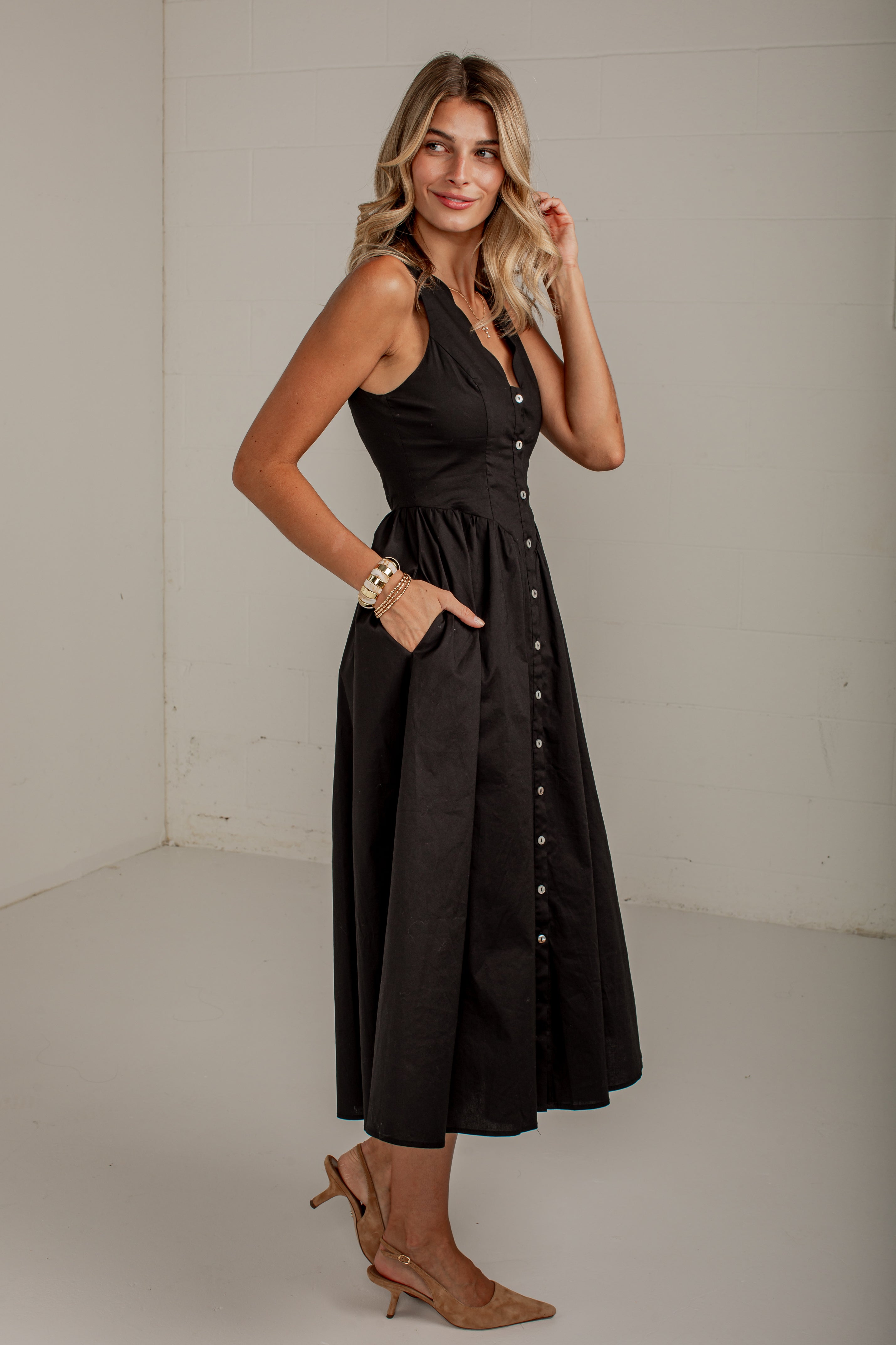 A woman modeling a sleeveless black midi dress with a buttoned front panel and side pockets, paired with tan heeled mules, in a plain studio setting.