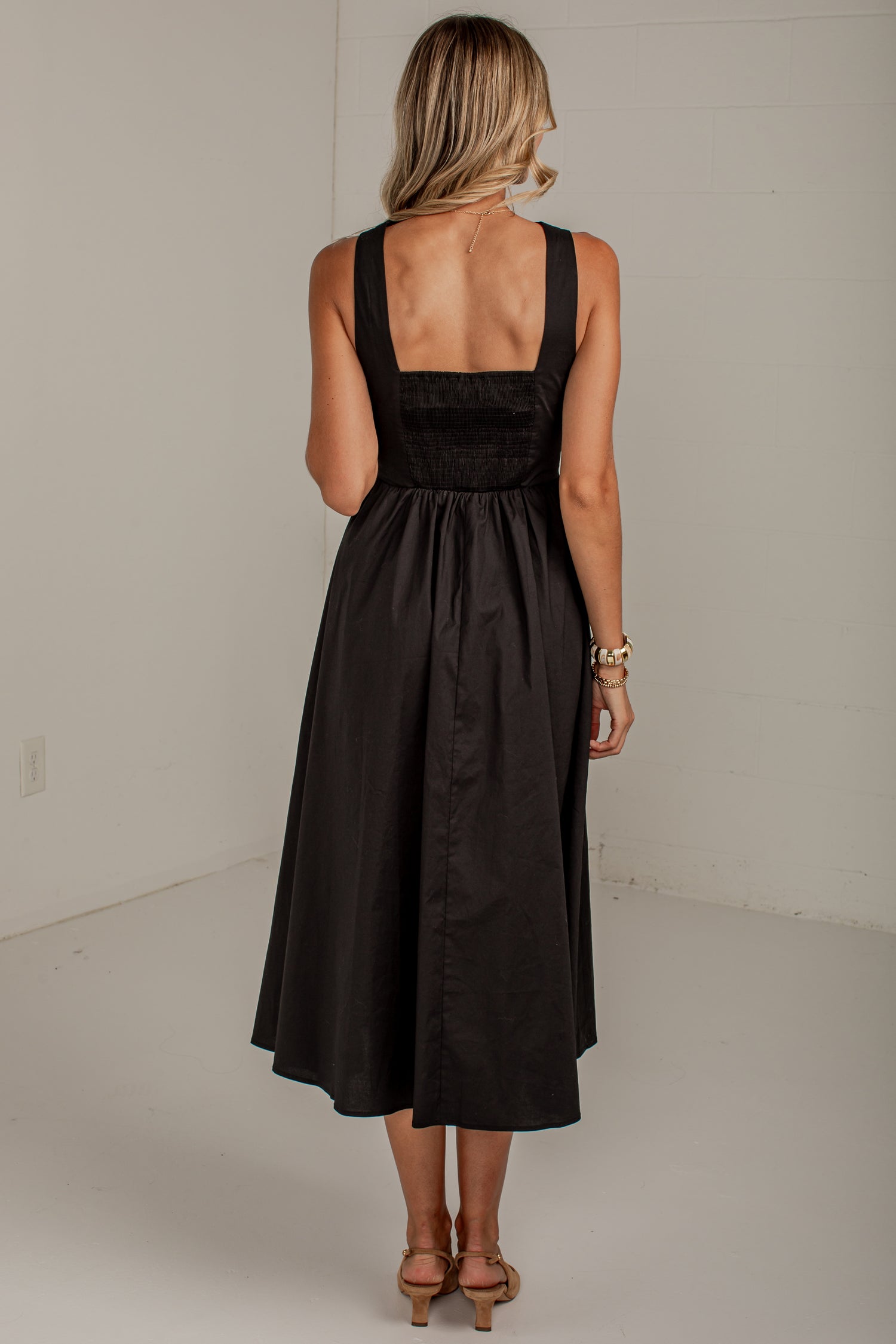 A woman viewed from behind wearing a black midi dress with a smocked bodice and wide shoulder straps, standing in a plain white studio setting.