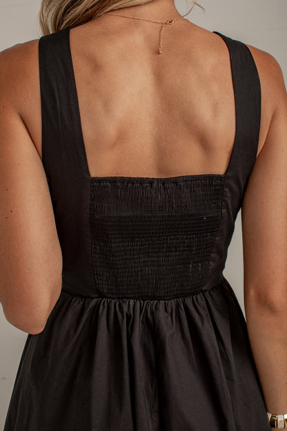 Close-up back view of a woman wearing a black sleeveless dress with a smocked bodice and wide straps against a plain light gray background.
