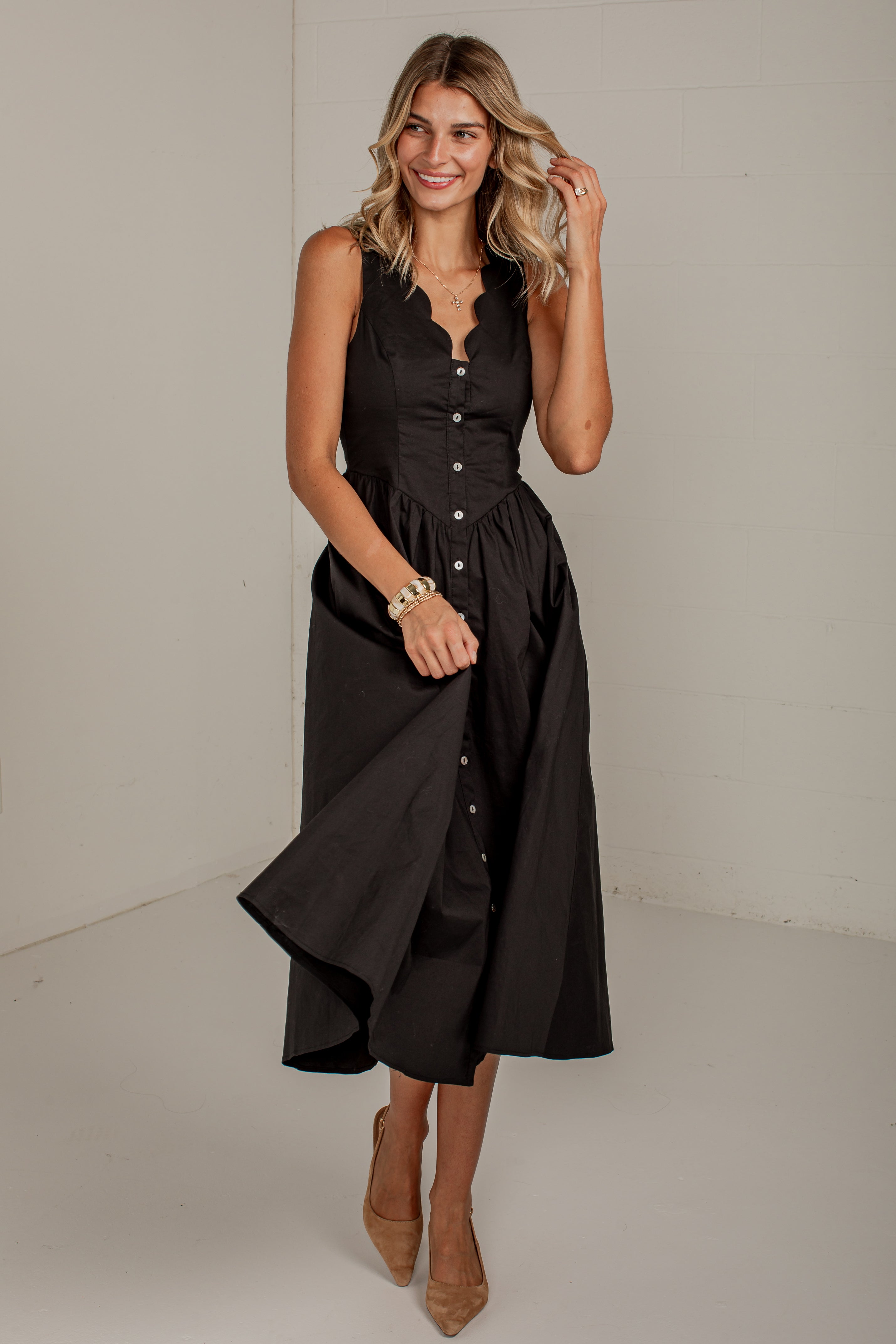 A woman modeling a sleeveless black midi dress with a button-front bodice and skirt, photographed in a plain indoor studio setting.