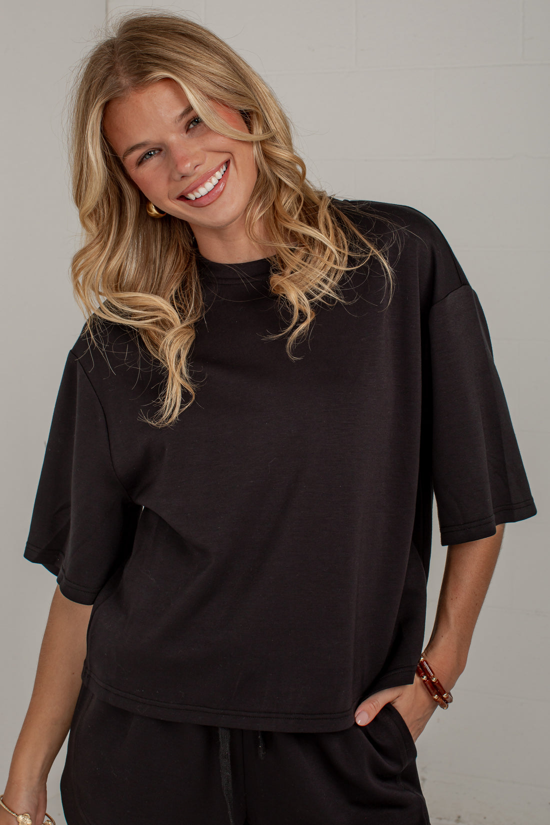 A smiling blonde woman wearing an oversized black short-sleeve t-shirt and matching black pants, standing against a light gray wall.