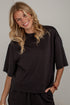A smiling blonde woman wearing an oversized black short-sleeve t-shirt and matching black pants, standing against a light gray wall.