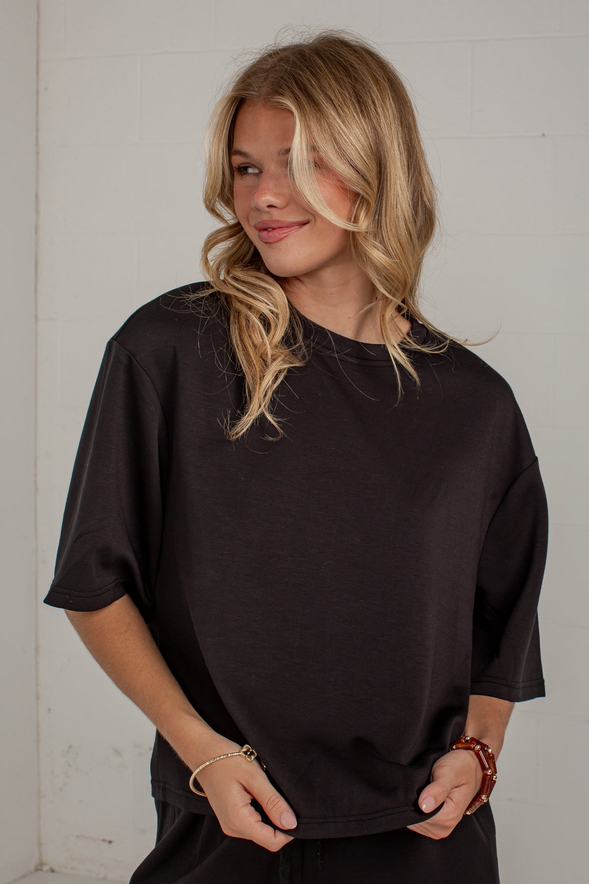 A woman with blonde hair wearing an oversized black short-sleeve t-shirt, standing against a white brick wall.
