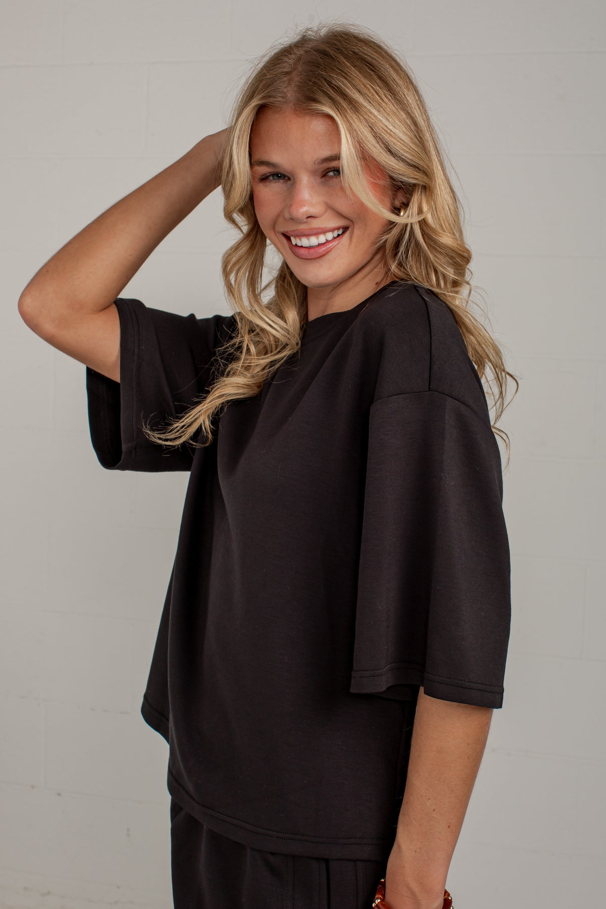 A smiling blonde woman wearing a loose black short-sleeve top and matching black bottoms, posing against a light gray wall.