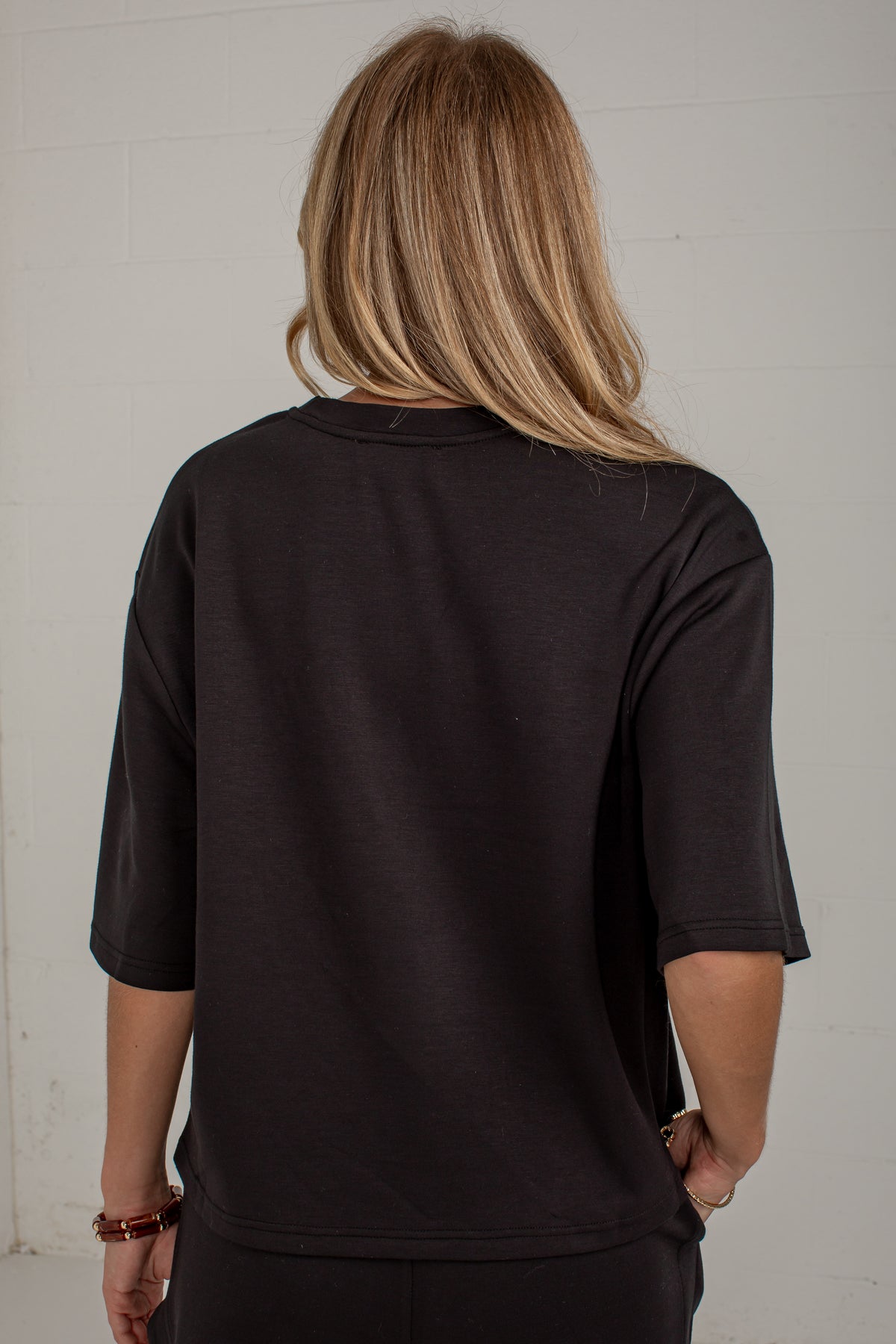 A woman viewed from behind wearing a black short-sleeve t-shirt and black pants, standing against a white brick wall.