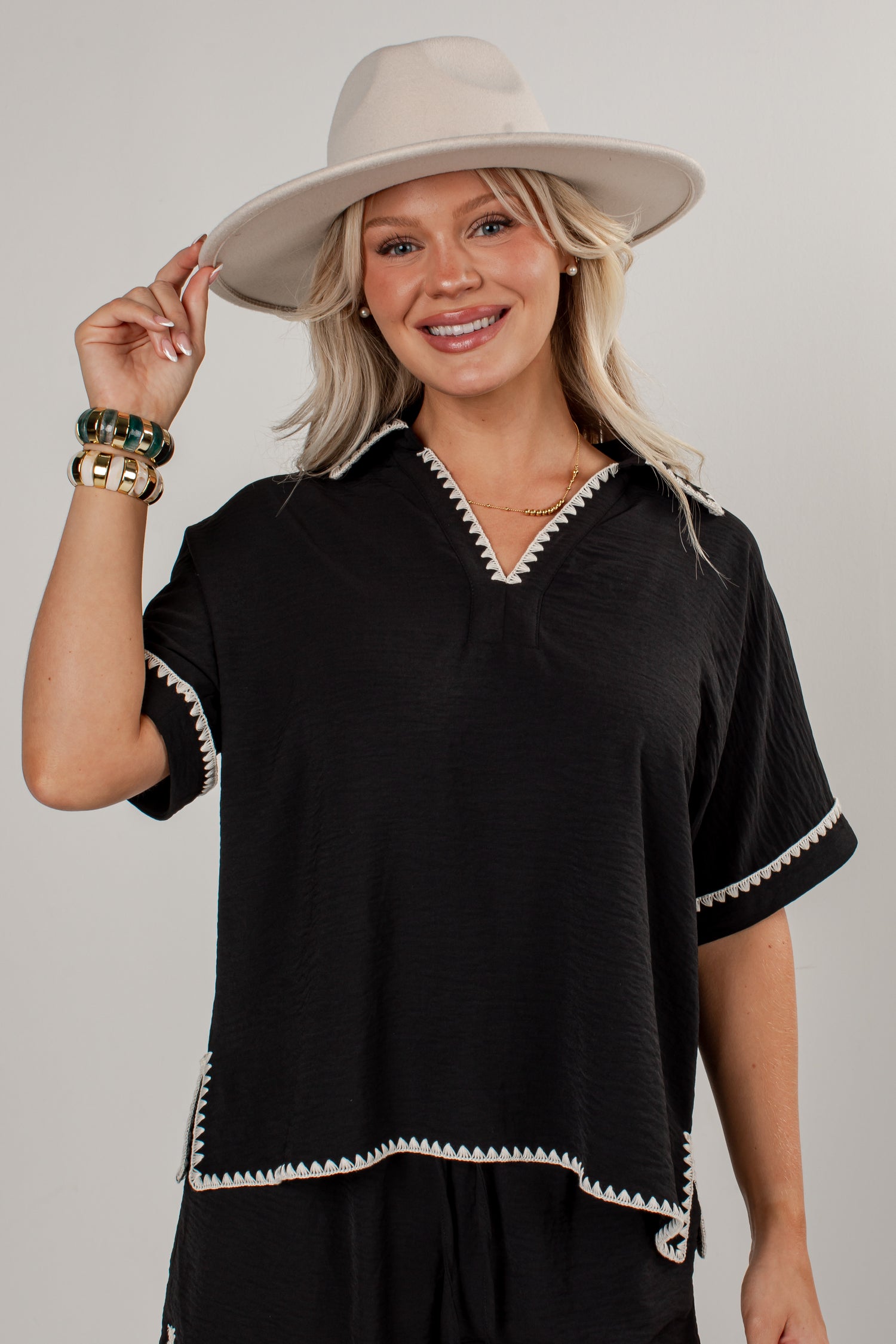 A woman wearing a black short-sleeve top with white geometric trim, a beige wide-brim hat, and gold bracelets, posing against a light gray studio background.