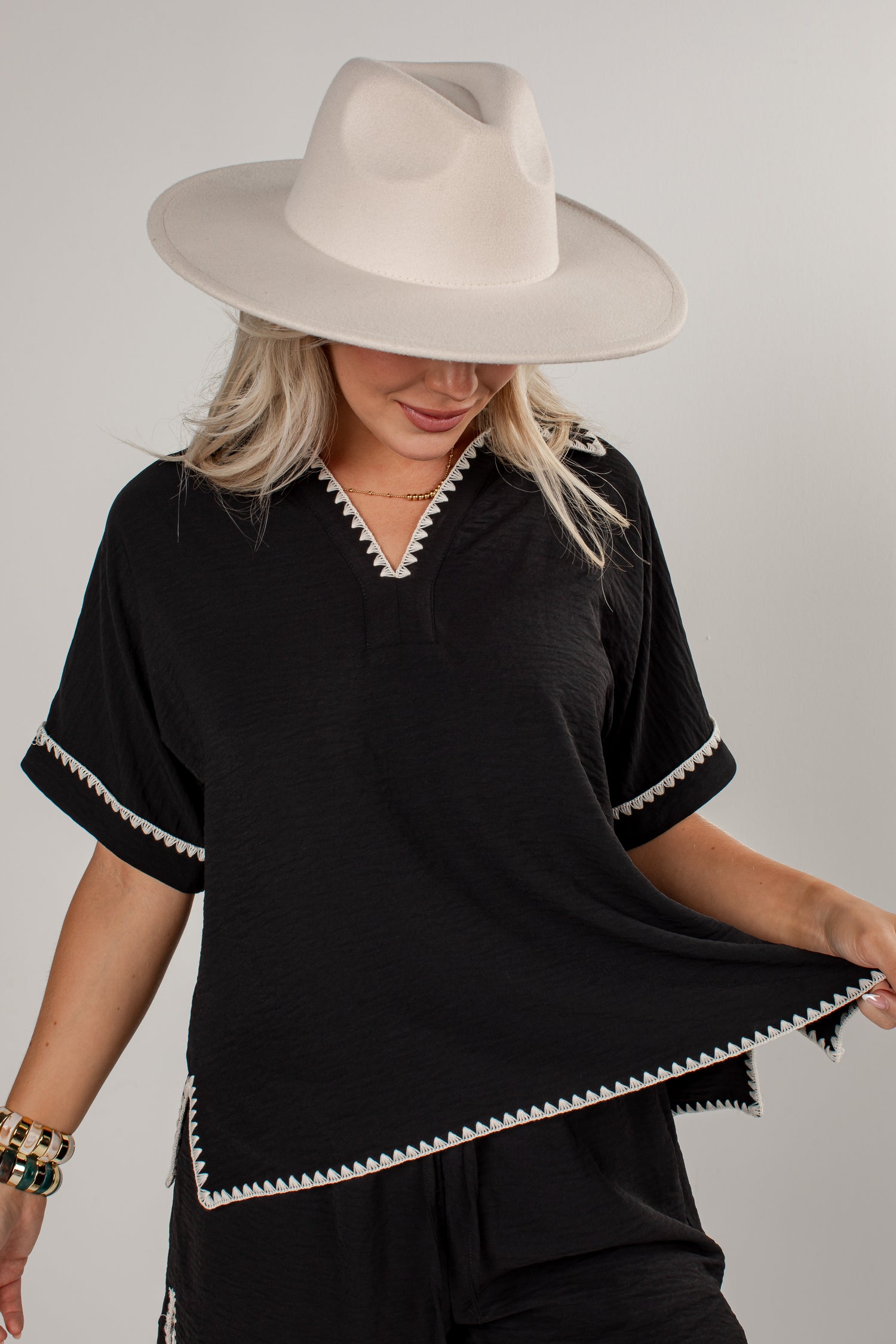 A woman wearing a black short-sleeve top with white decorative trim along the V-neck, sleeve cuffs, and hem, paired with a wide-brimmed cream hat against a light gray studio background.