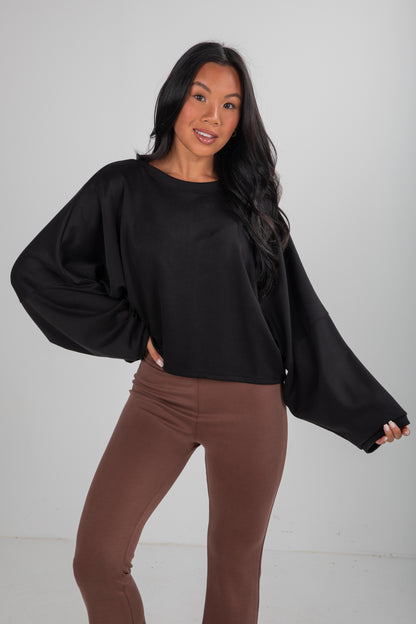 Woman wearing a black top with wide sleeves and brown pants on a plain background