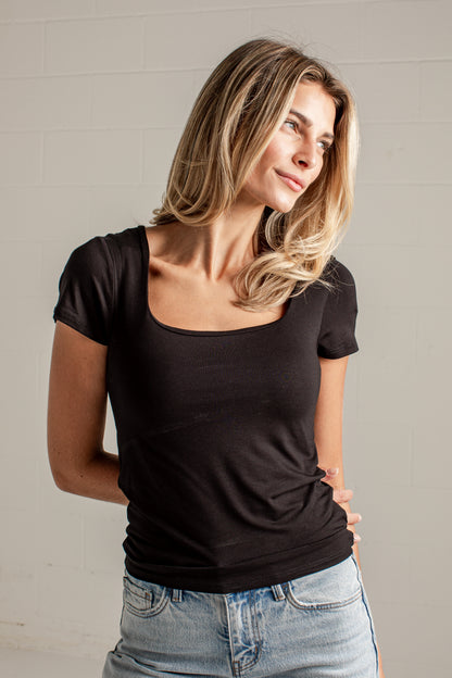 A woman wearing a black scoop-neck short-sleeve t-shirt and light-wash blue jeans, standing against a light gray brick wall.