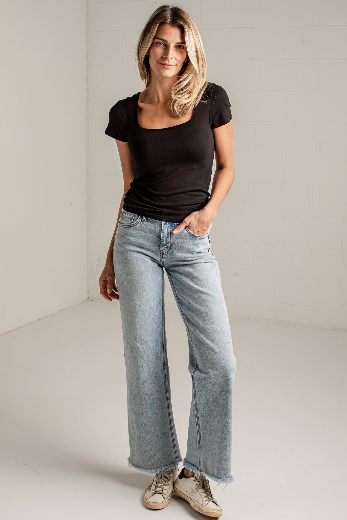 A woman modeling a black scoop-neck short-sleeve t-shirt and light-wash wide-leg jeans, standing in a bright white studio corner.