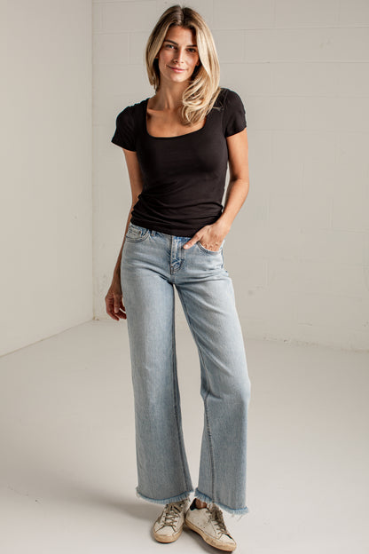 A woman modeling a black scoop-neck short-sleeve t-shirt and light-wash wide-leg jeans, standing in a bright white studio corner.