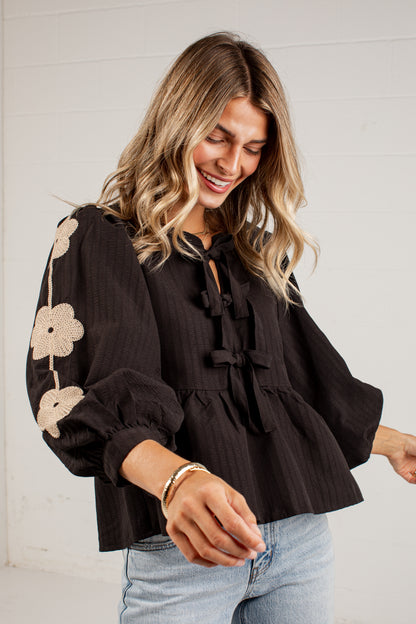 A woman wearing a black long-sleeve blouse with beige floral embroidery on the sleeve and tie-front bow details, paired with light blue jeans, standing against a white paneled wall.