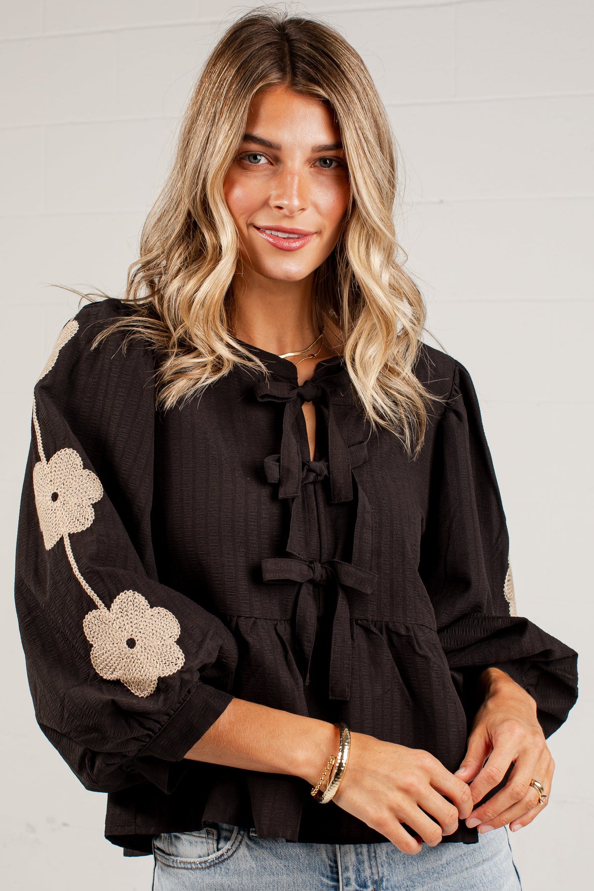 A woman wearing a black long-sleeve blouse with beige floral embroidery on the sleeve and tie-front detailing, paired with light-wash blue jeans, standing against a white paneled wall.
