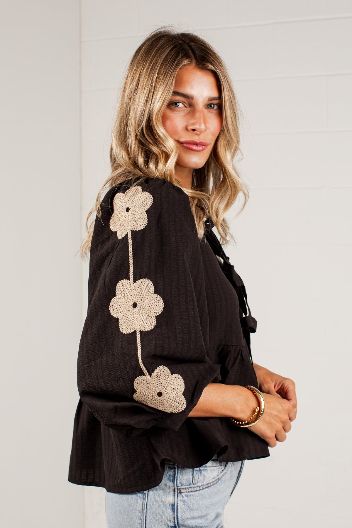 A woman wearing a black long-sleeve blouse with beige crochet flower appliqués down the sleeve, paired with light blue jeans, standing against a white paneled wall.