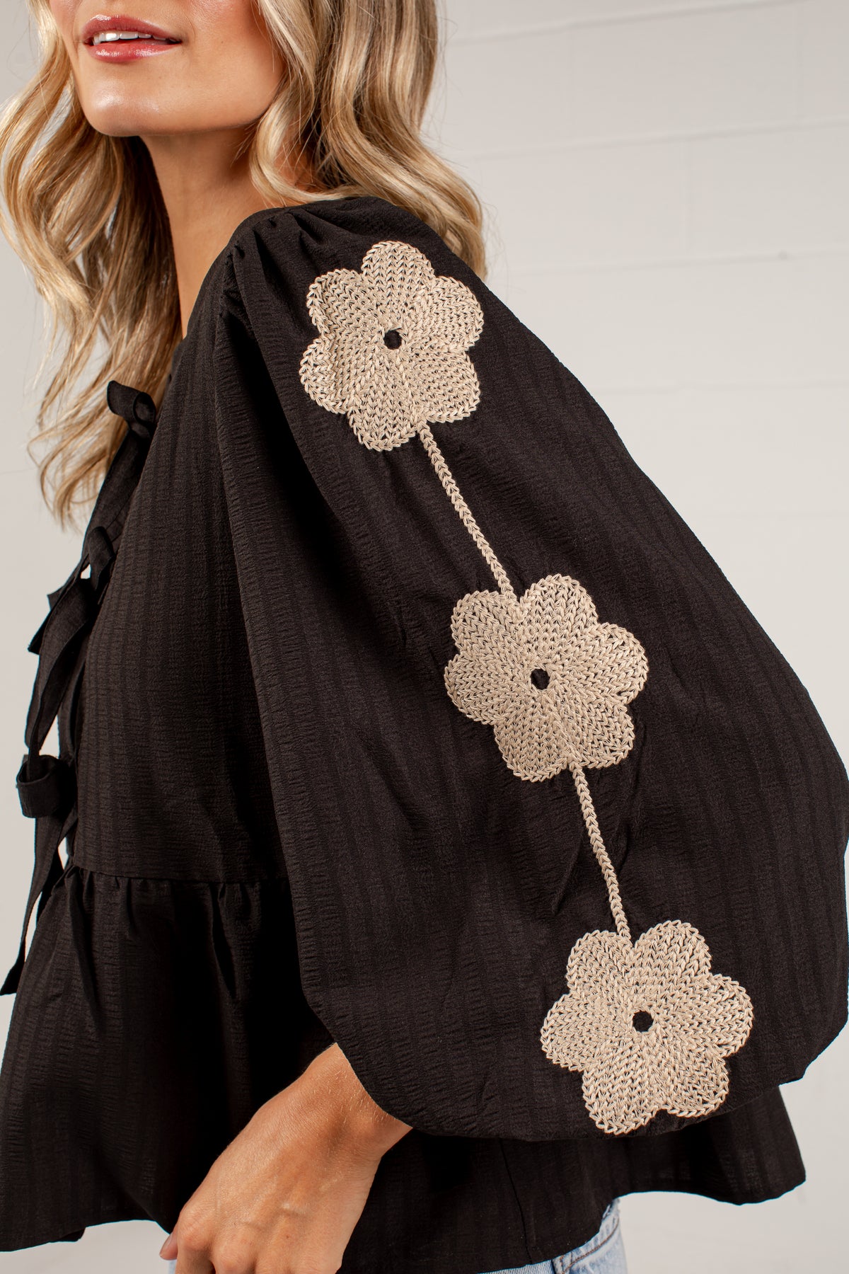 A close-up of a woman wearing a black textured blouse with three large beige crochet flower appliqués down one sleeve, paired with light blue jeans against a white paneled wall.